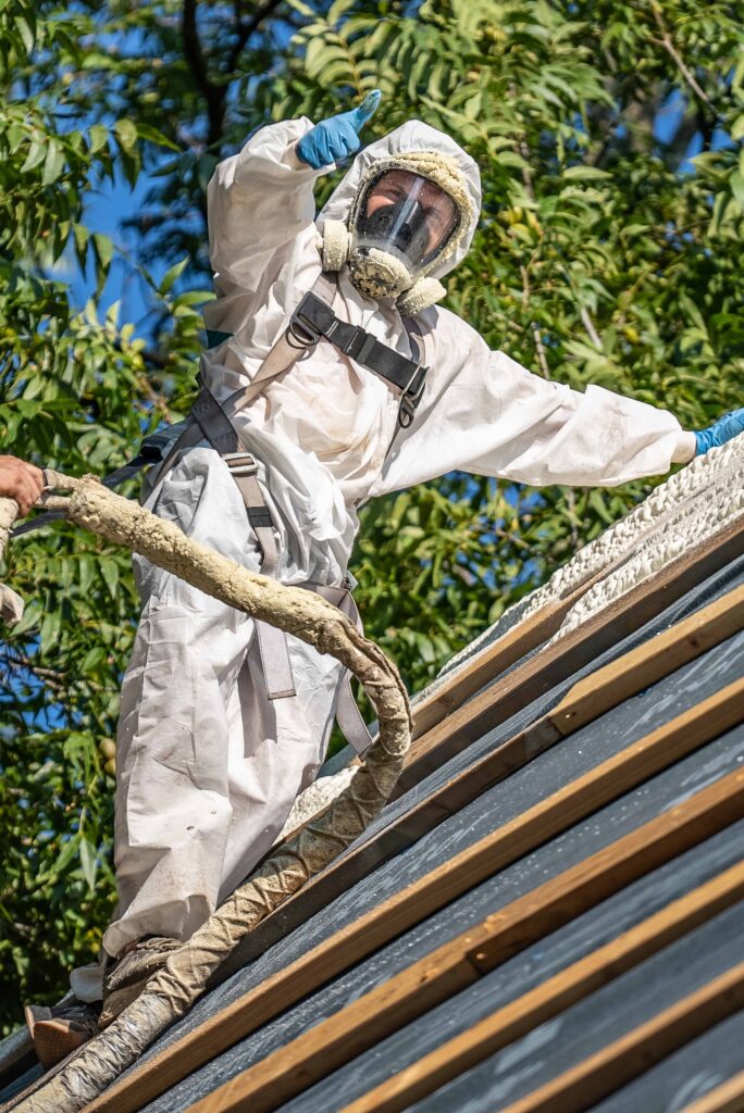 A worker in protective gear, including a mask and gloves, is skillfully applying Arrow Insulations spray foam on a rooftop. They balance on a ladder with a long hose in hand, surrounded by lush green foliage under the bright sunny sky.