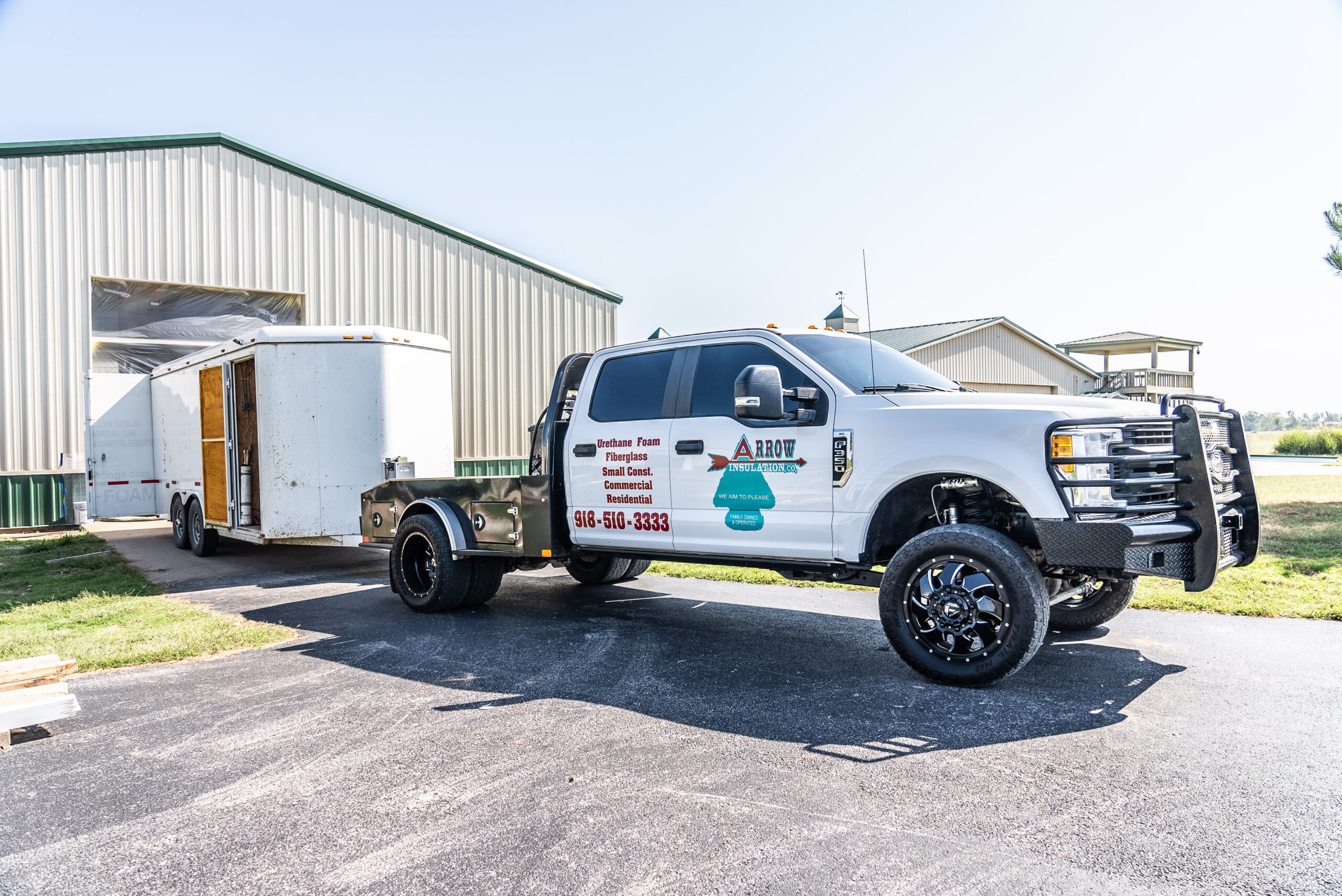 A white truck with Arrow Insulation branding is parked on a paved area in front of a large metal warehouse. Hitched to the truck is a white enclosed trailer. Sunlight casts shadows on the ground, highlighting their focus on spray foam insulation solutions.