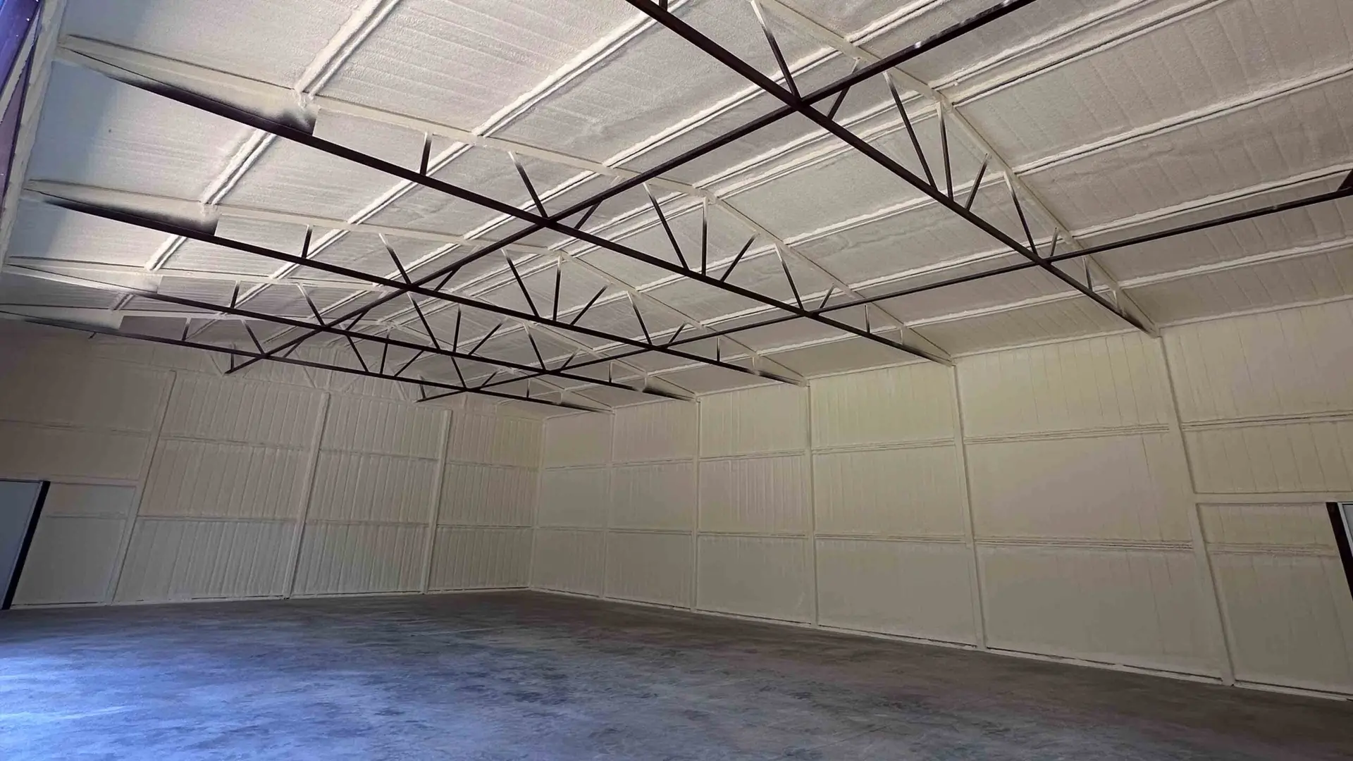 Large empty warehouse with white Arrow Insulation walls and ceiling, exposed steel roof trusses, and a concrete floor. Natural light enters from a partially open door on the left side.